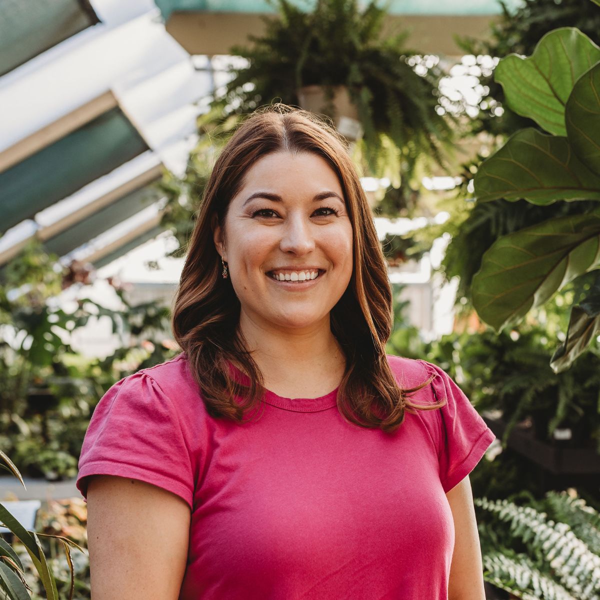 Headshot of Maria in a pink dress standing in a greenhouse with greenery behind her
